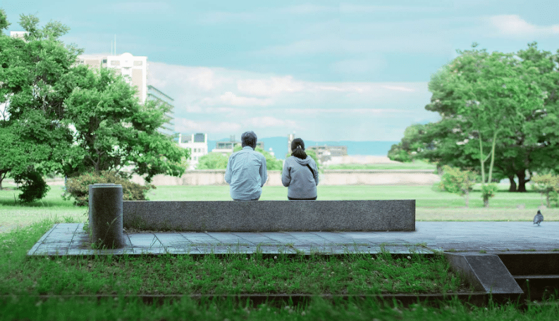 Couple sat on a concrete bench looking out over a park with buildings in the distance